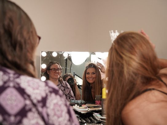 Kellie Lightbourn smiling in a backstage mirror as a woman is crowned