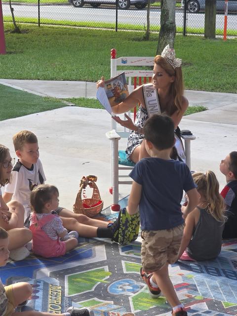 Kellie Lightbourn, in crown and sash, reading a picture book to young children outdoors