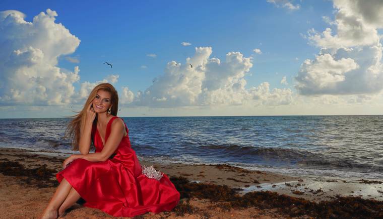 Kellie Lightbourn, Mrs. Universe® 2026, smiling, seated on the sand in a red gown with the crown beside her
